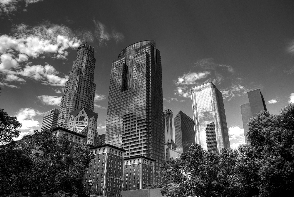 black and white photo of downtown Los Angeles' skyscrapers, with some clouds in the sky reflected back in an imposing manner.