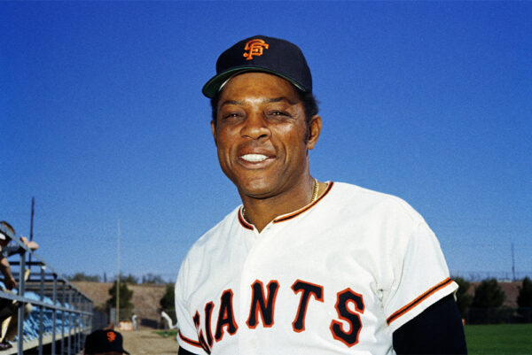 Willie Mays smiles at the camera. He wears a black cap with the letters "SF" and a white baseball uniform with the large letters "Giants."