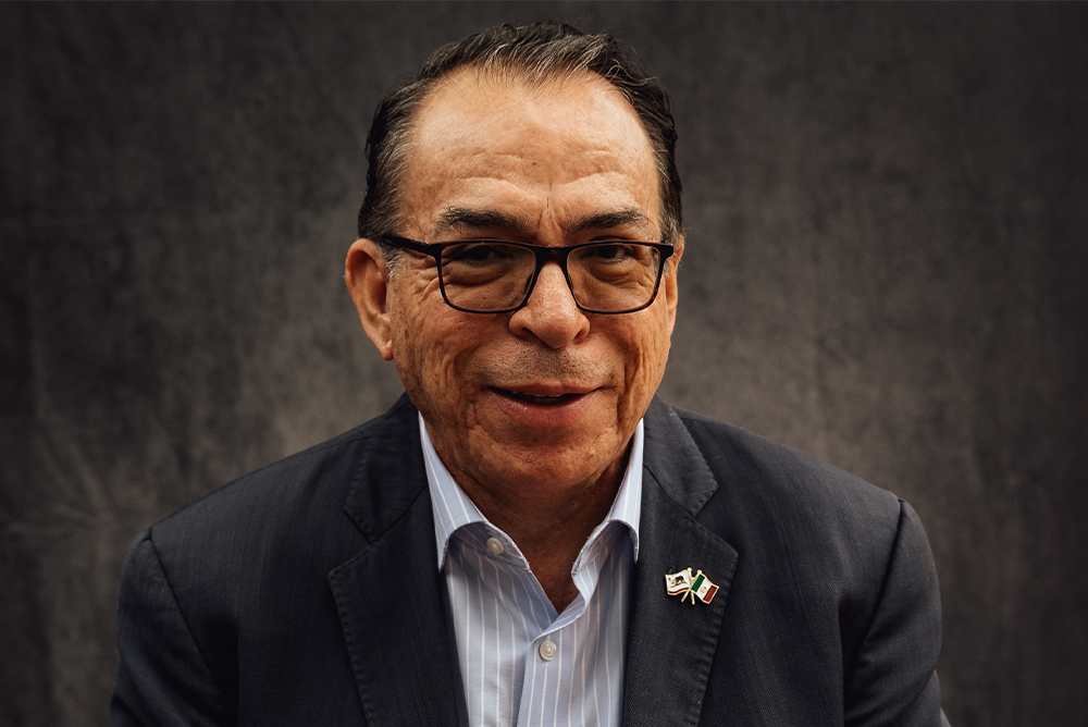 A headshot of Alfredo Corchado smiling against a gray-black backdrop. He wears black-rimmed glasses and a black jacket on top of a blue collared shirt. A small pin of California's official state flag and Mexico's official flag waving is pinned to right side of his jacket.