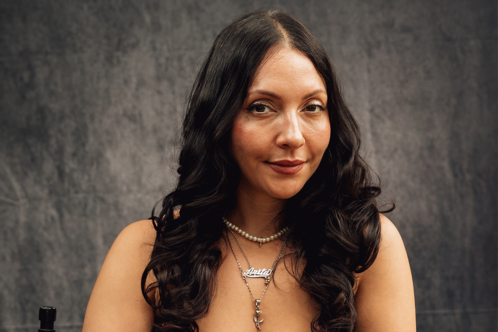 A headshot of Anita Herrera smiling against a gray-black backdrop. Her face is slightly turned to the right. A pearl necklace is around her neck. She also wears a necklace that says, "Anita."