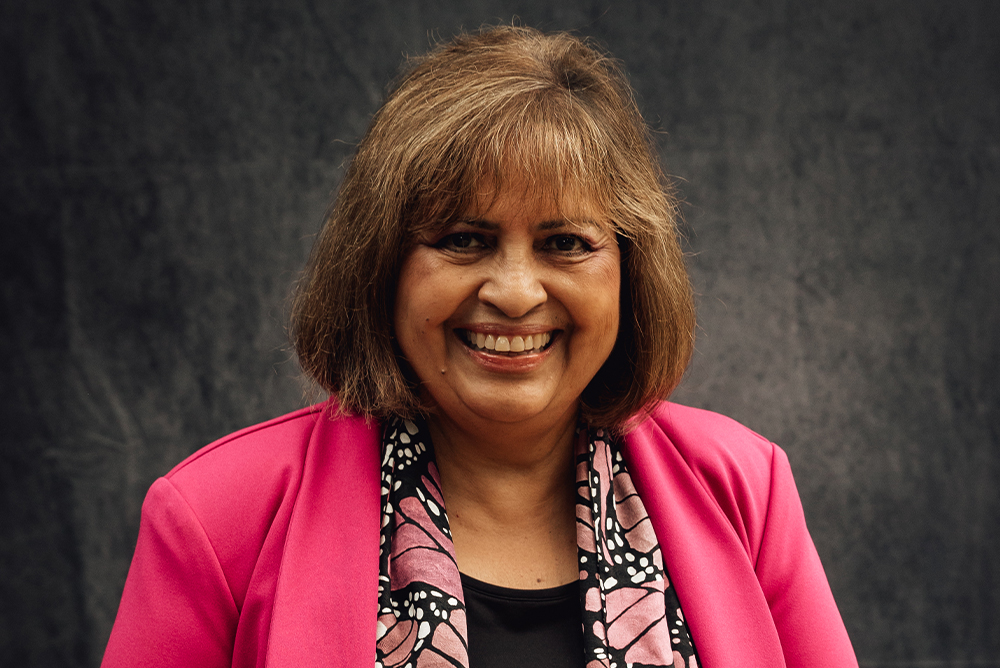 A headshot of Irasema Coronado smiling against a gray-black backdrop. She wears a pink jacket over a black shirt with a colorful scarf.